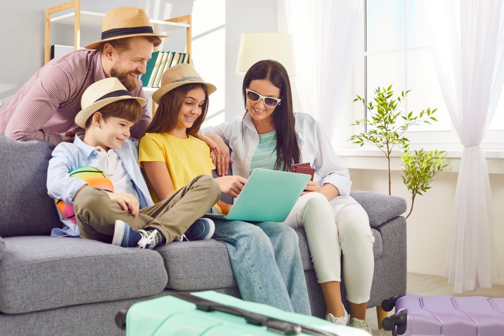 Young happy family booking tickets online via laptop in internet sitting on sofa in living room at home together. Parents with kids waiting for summer vacation trip. Travel concept.